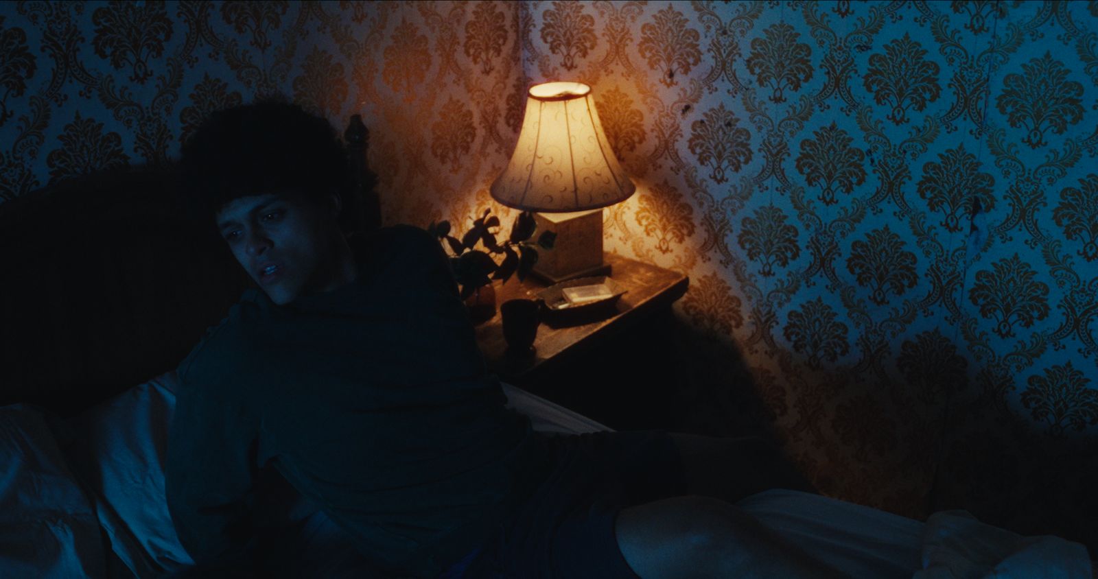 A bedside lamp in a wallpapered room, from Sundered